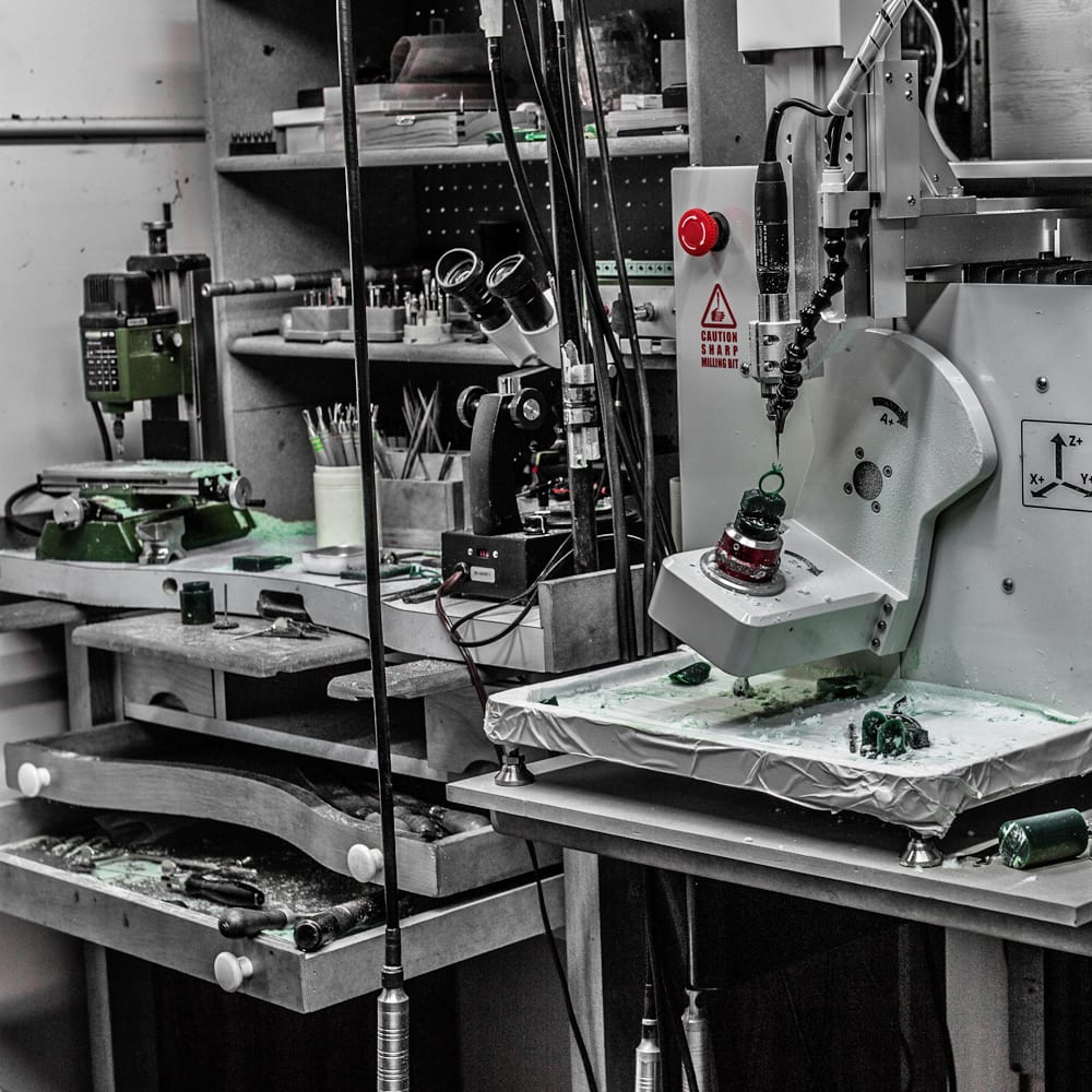 A desktop 4-axis milling machine cutting green jewelry wax beside a jeweler's bench and microscope