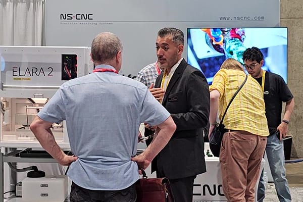 An NS CNC team member talking with visitors at the NS CNC trade show booth, beside an Elara 2