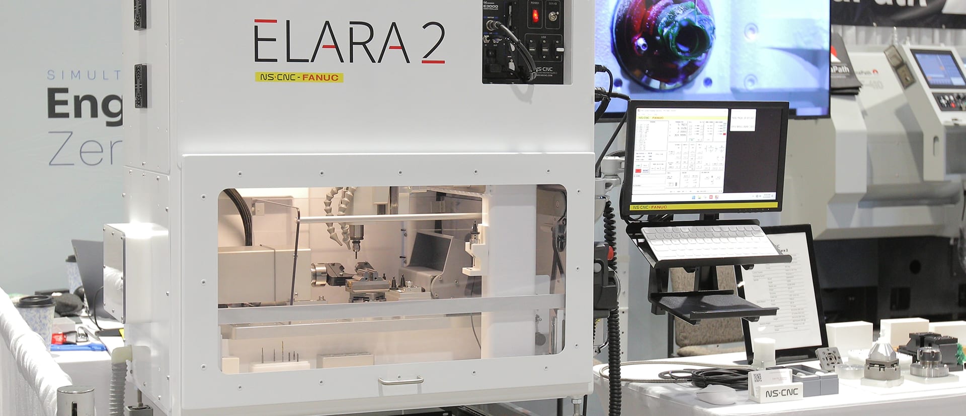 An Elara 2 with NS CNC-FANUC controls on display at a trade show, beside its control monitor and a table of machined parts