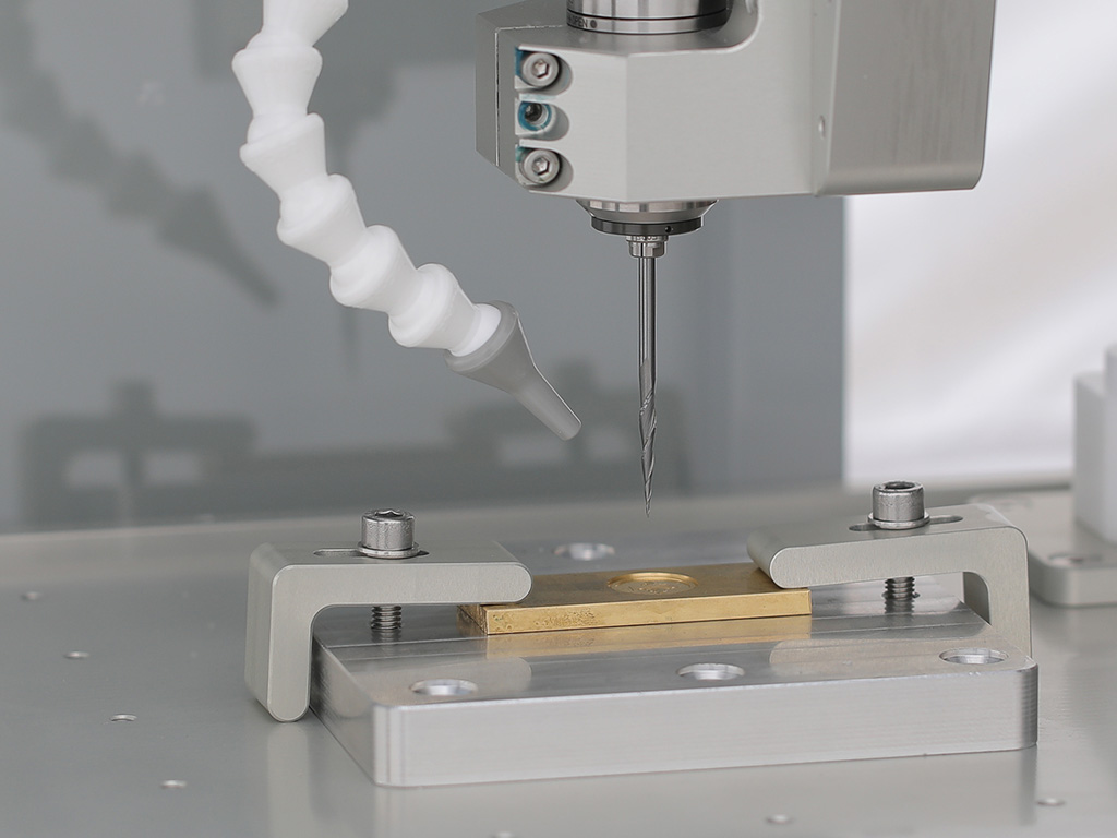 Thin brass plate held by two low-profile edge clamps on a fixture plate, tapered engraving cutter above