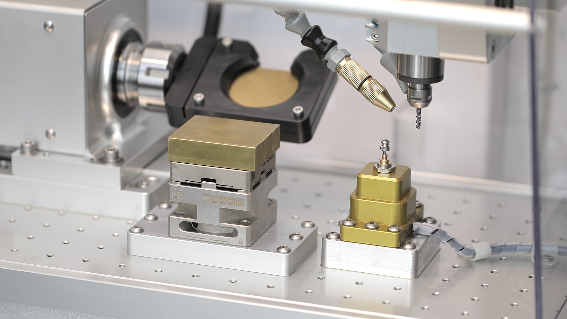 Inside the Elara 2 work area: rotary axis with trunnion plate, stacked pallet vise with brass block, and a gold stepped quick-release fixture under the drill